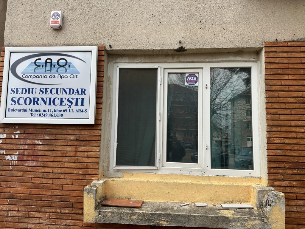 cao scornicesti WhatsApp Image 2026-08-14 at 145708 - Jurnalul Olteniei – Știri din Craiova și Oltenia Storefront with a large blue and white sign on a brick wall, beside a glass door and a small alarm box above.
