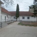 White single-story building with a red-tiled roof, a tall evergreen tree in the courtyard, and a paved walkway.