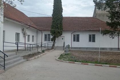 White single-story building with a red-tiled roof, a tall evergreen tree in the courtyard, and a paved walkway.