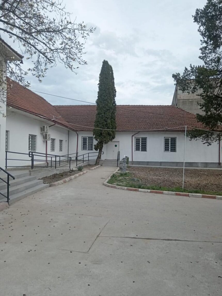 White single-story building with a red-tiled roof, a tall evergreen tree in the courtyard, and a paved walkway.