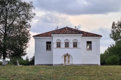 Small white chapel with a red tile roof and central arched doorway, flanked by three small arched windows, in a grassy field.