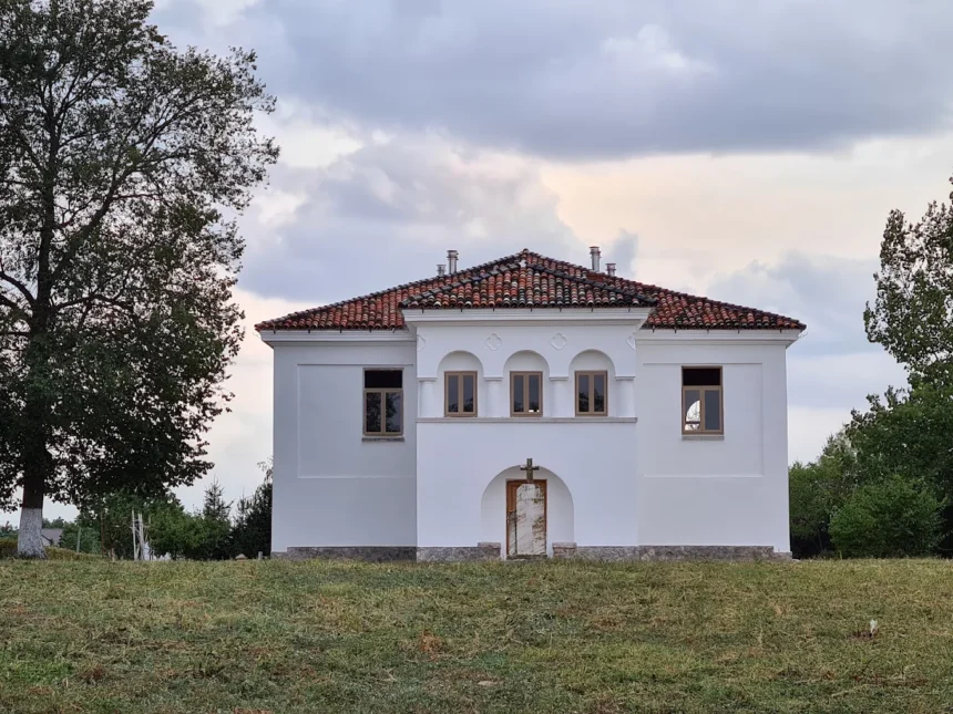 Small white chapel with a red tile roof and central arched doorway, flanked by three small arched windows, in a grassy field.