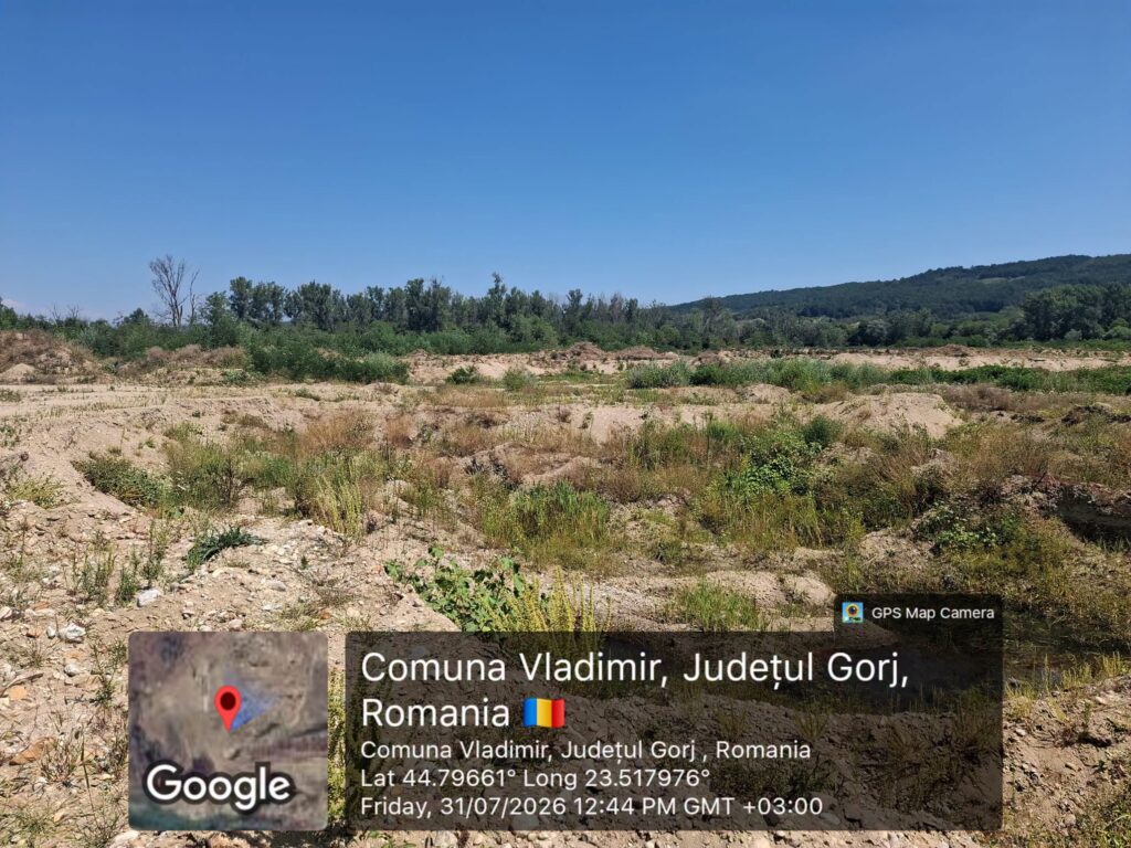 Wide view of a dry, rocky field with sparse shrubs under a clear blue sky and a distant tree line/hills on the horizon; overlaid location text shows Comuna Vladimir, Judetul Gorj, Romania.