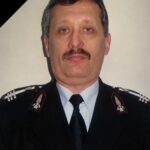 Portrait of a male police officer in dress uniform with a mourning black ribbon across the top-left corner, badge visible on chest.