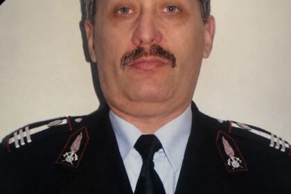 Portrait of a male police officer in dress uniform with a mourning black ribbon across the top-left corner, badge visible on chest.