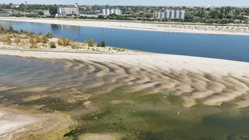 Wide river with exposed sandbars and green algae near the shore; industrial buildings and silos visible in the background.
