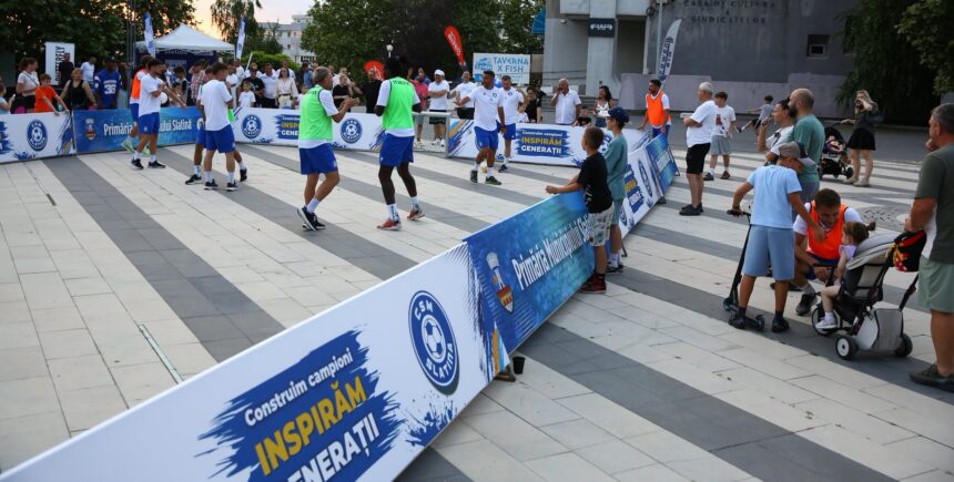 Group of players in a small outdoor futsal court, spectators along the sidelines with blue advertising banners in Romanian.