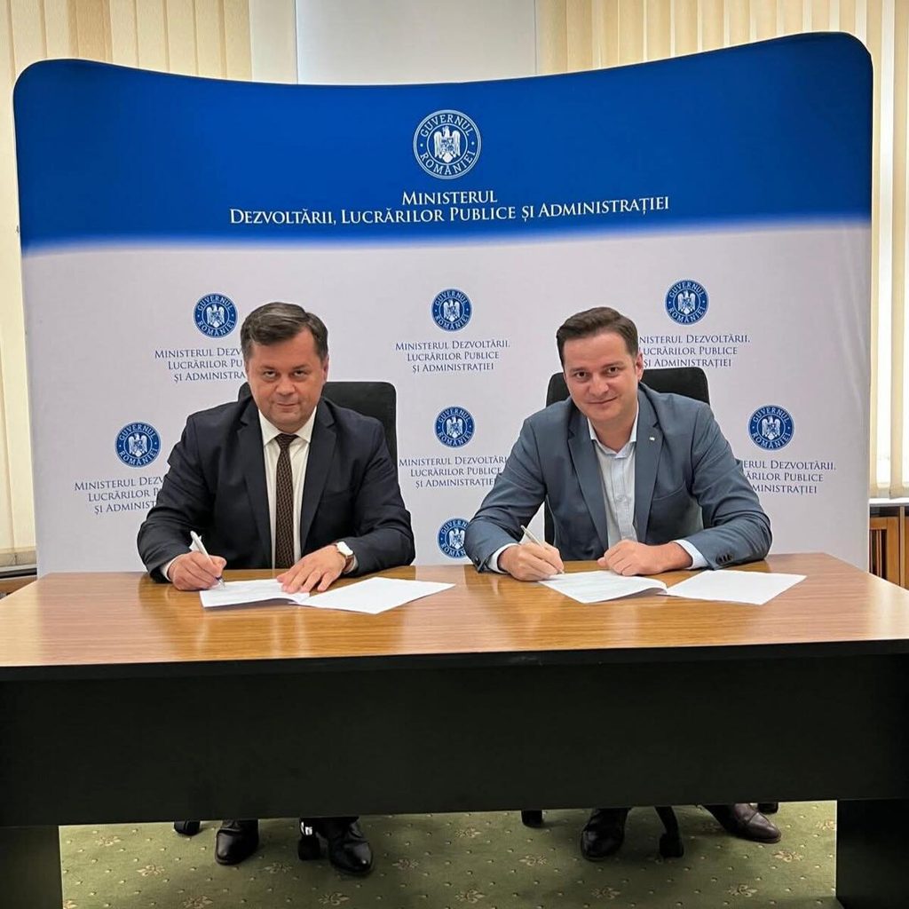 Two men in suits sign documents at a wooden table during a formal agreement, with a blue-and-white ministry backdrop behind them (Romania).
