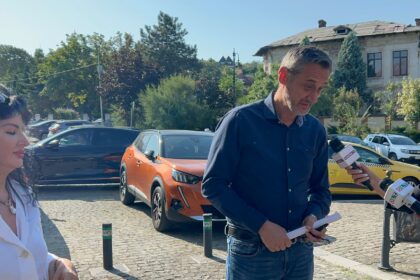 Man in a blue shirt speaks to reporters on a sunny street, holding notes while microphones are waved toward him, with a woman on the left and cars in the background.