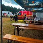 Outdoor emergency response scene under a blue canopy with wooden benches; a red ambulance and responders behind yellow caution tape in a grassy field.