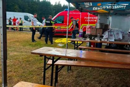 Outdoor emergency response scene under a blue canopy with wooden benches; a red ambulance and responders behind yellow caution tape in a grassy field.