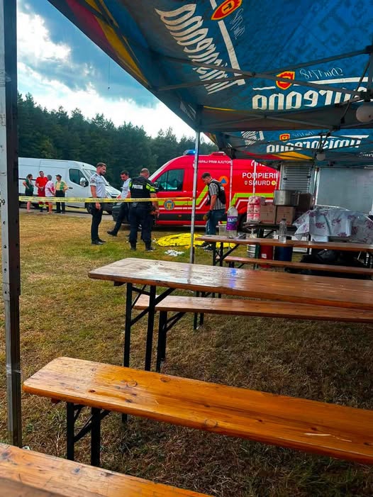 Outdoor emergency response scene under a blue canopy with wooden benches; a red ambulance and responders behind yellow caution tape in a grassy field.