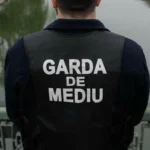 Back view of a person wearing a dark vest that reads GARDA DE MEDIU, standing on a bridge over water.
