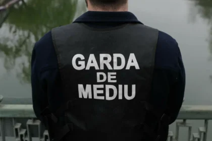 Back view of a person wearing a dark vest that reads GARDA DE MEDIU, standing on a bridge over water.