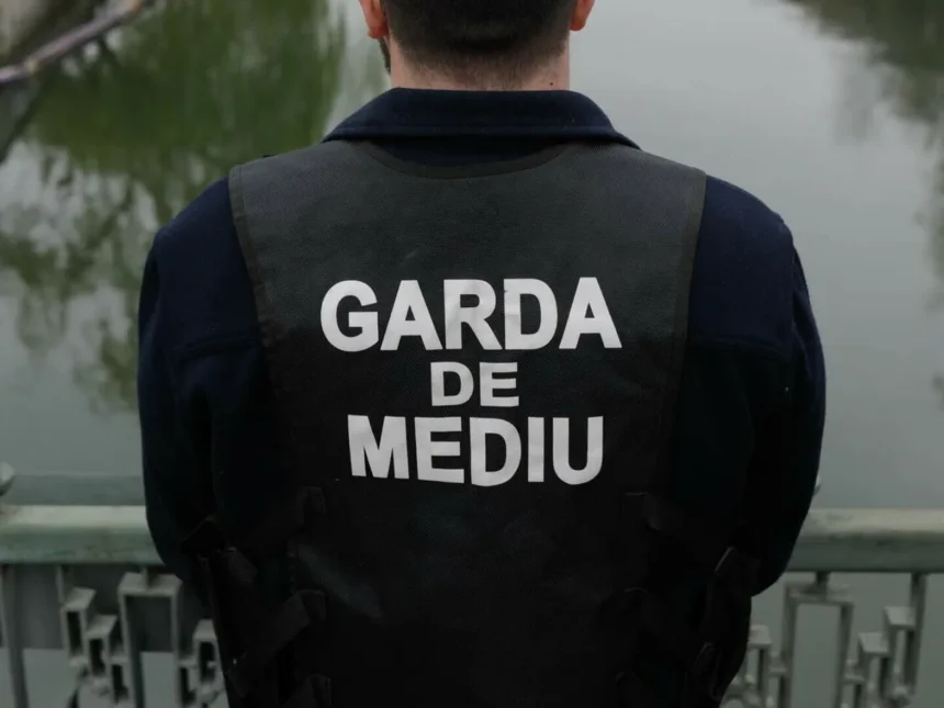 Back view of a person wearing a dark vest that reads GARDA DE MEDIU, standing on a bridge over water.