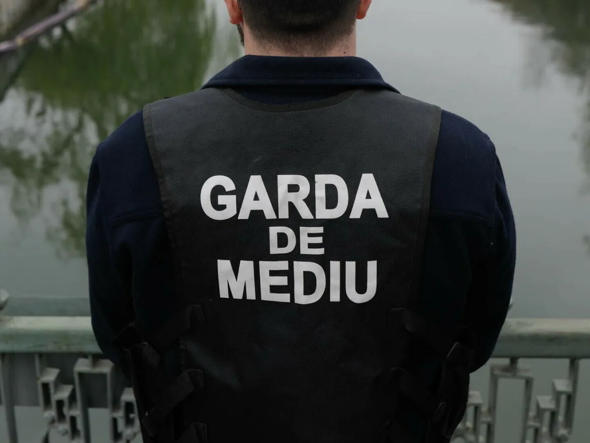 garda de mediu dONj2pEiSifA79A5kjnLt7IYs8pWjh7XhufSKEr9 - Jurnalul Olteniei – Știri din Craiova și Oltenia Back view of a person wearing a dark vest that reads GARDA DE MEDIU, standing on a bridge over water.