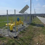 Yellow pipeline valves and meters fenced along a rural roadside with solar panel on a pole and open fields nearby
