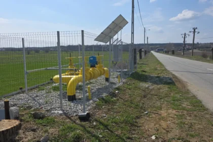 Yellow pipeline valves and meters fenced along a rural roadside with solar panel on a pole and open fields nearby