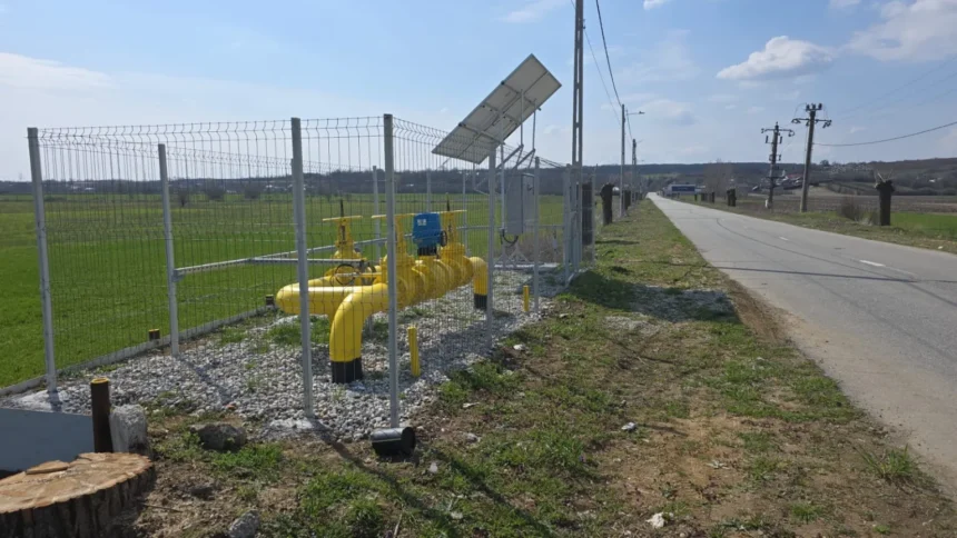 Yellow pipeline valves and meters fenced along a rural roadside with solar panel on a pole and open fields nearby