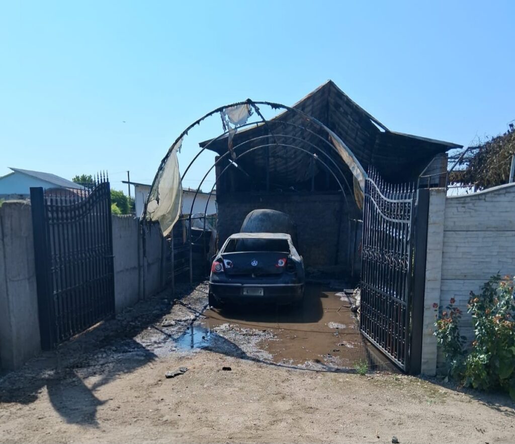 Black VW sedan parked in a muddy driveway under a burnt, half-collapsed carport with torn fabric; iron gates stand open on both sides.