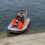 Inflatable rescue boat with four crew members in orange life jackets near a concrete shore; officer at the console with a red-blue light bar overhead.