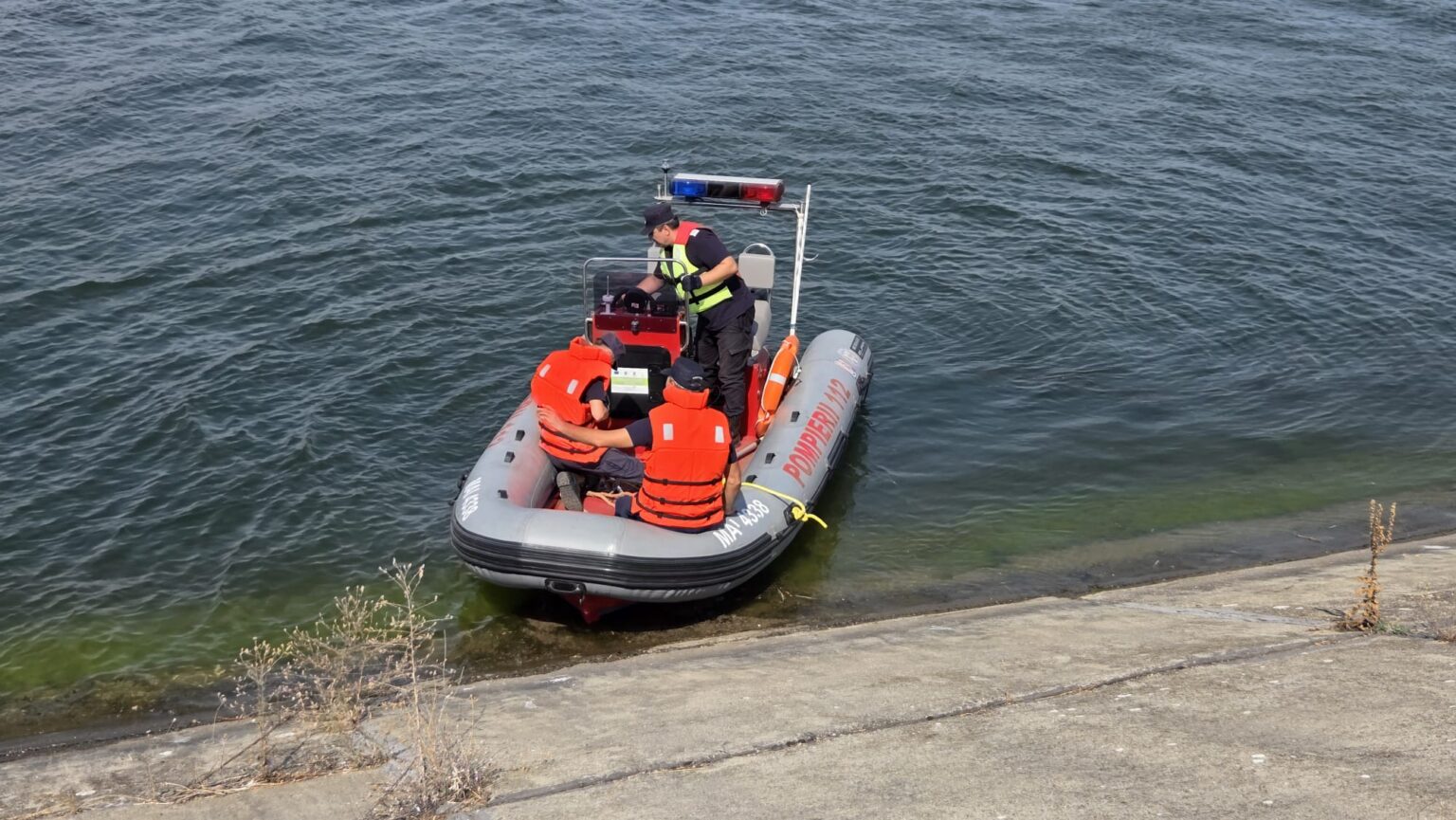 Inflatable rescue boat with four crew members in orange life jackets near a concrete shore; officer at the console with a red-blue light bar overhead.