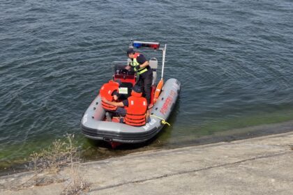 Inflatable rescue boat with four crew members in orange life jackets near a concrete shore; officer at the console with a red-blue light bar overhead.