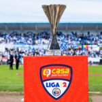 Silver trophy on a bright red podium with a Liga 12 crest at the front; blurred stadium crowd in the background.