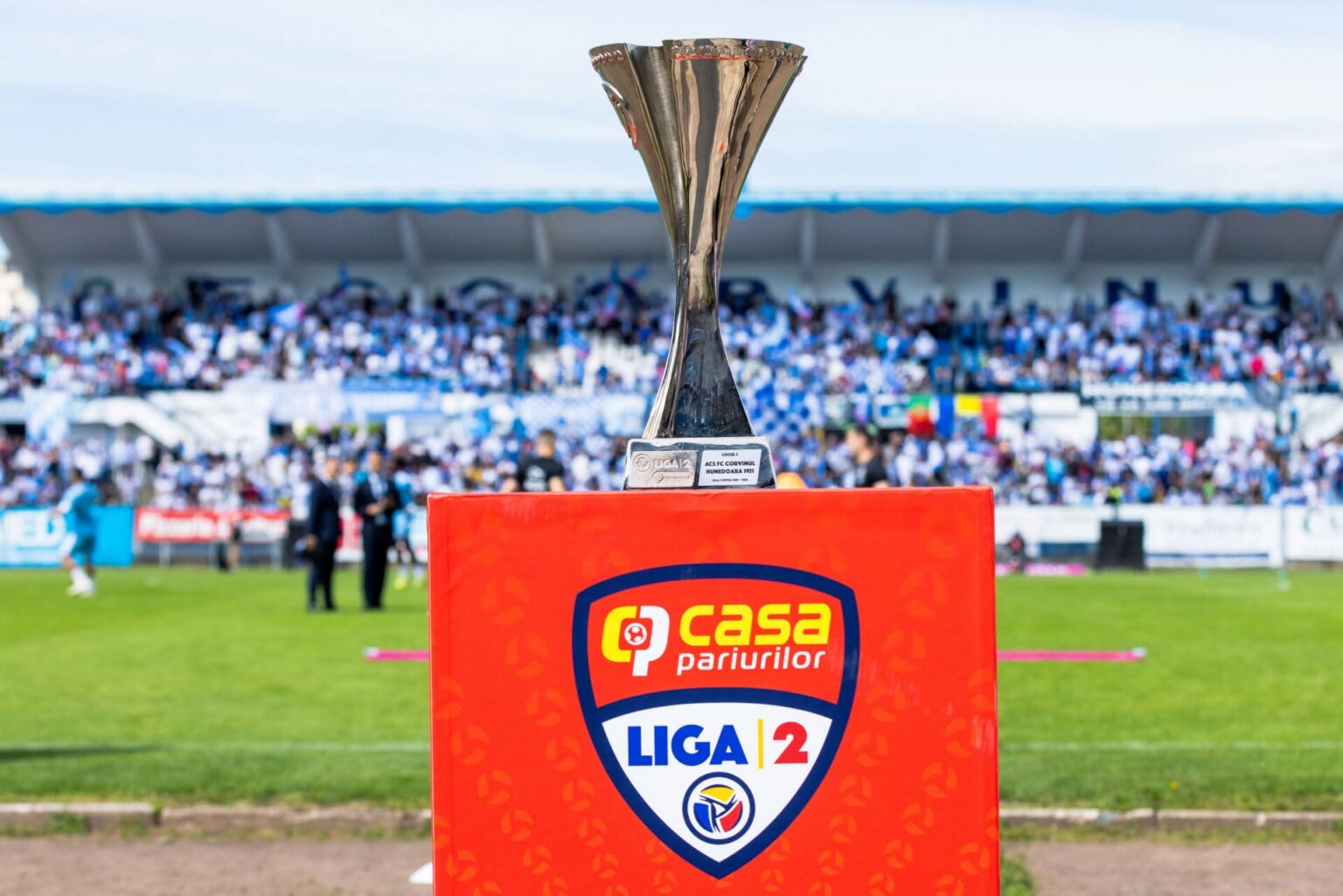 Silver trophy on a bright red podium with a Liga 12 crest at the front; blurred stadium crowd in the background.