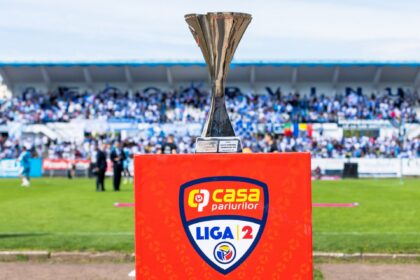 Silver trophy on a bright red podium with a Liga 12 crest at the front; blurred stadium crowd in the background.