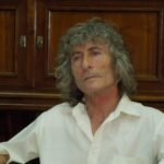 Portrait of an older woman with gray curly hair, wearing a white shirt, seated indoors with dark wood cabinets behind her.