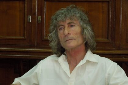 Portrait of an older woman with gray curly hair, wearing a white shirt, seated indoors with dark wood cabinets behind her.