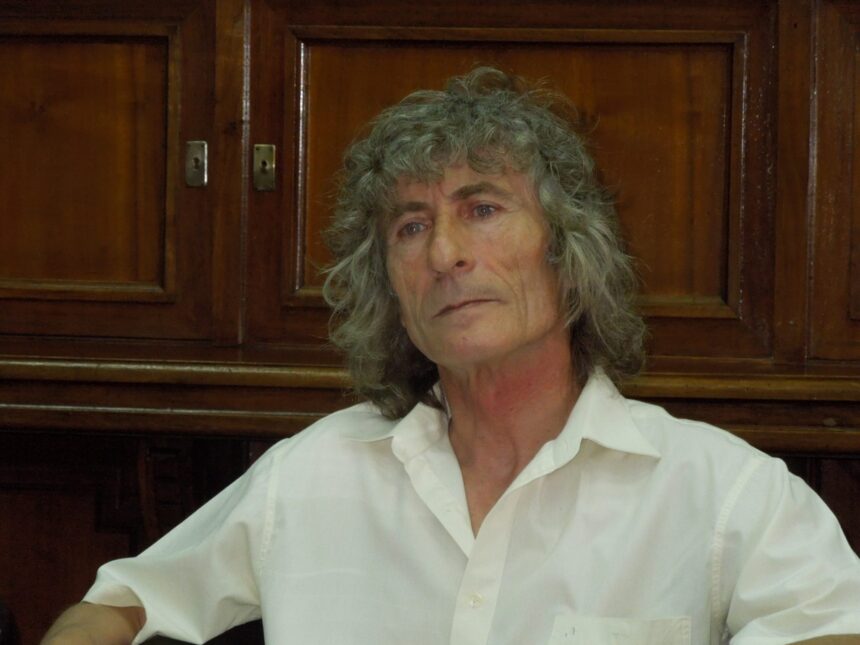 Portrait of an older woman with gray curly hair, wearing a white shirt, seated indoors with dark wood cabinets behind her.