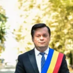 Man in a dark suit and blue tie wearing a blue-yellow-red sash, standing outdoors in a sunlit park.