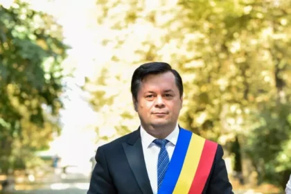 Man in a dark suit and blue tie wearing a blue-yellow-red sash, standing outdoors in a sunlit park.