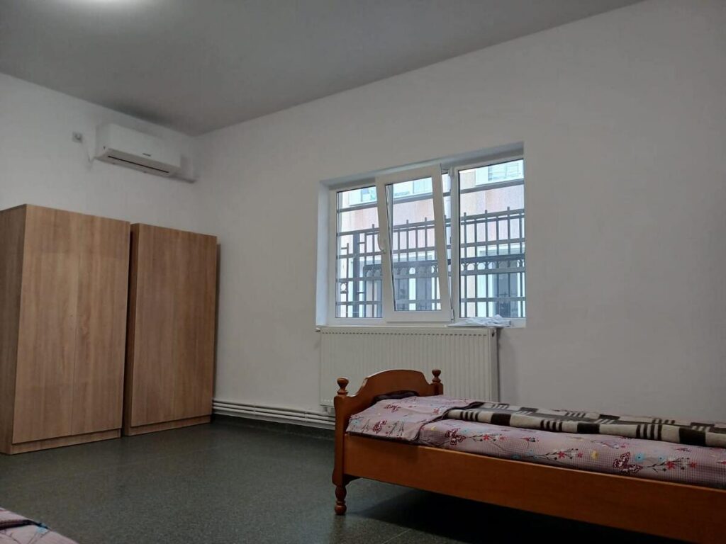 Sparse bedroom with two tall wooden wardrobes, a single bed with a floral blanket, window with bars, and a wall heater.