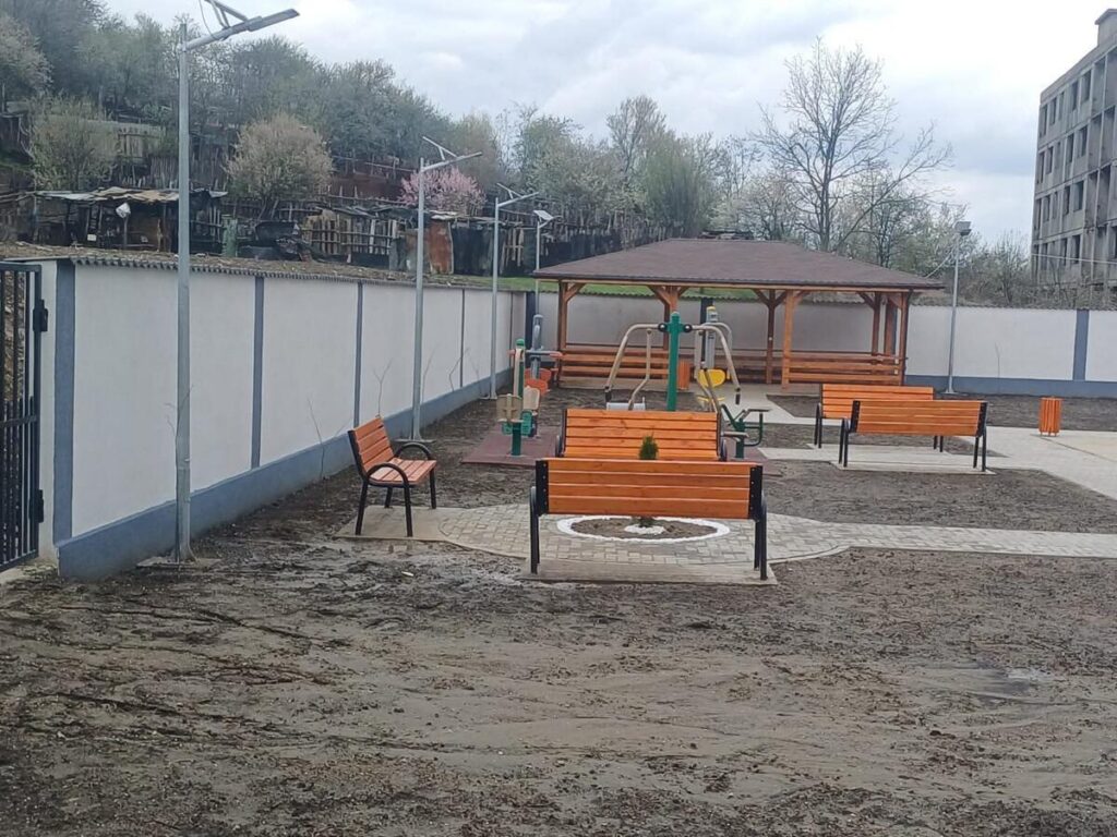 Public park area with wooden benches around a small playground and a wooden pavilion; muddy ground and bare trees under an overcast sky.