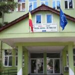 Green school building with a covered entrance, Romanian and EU flags on the roof, and a sign identifying Colegiul National Ion Minulescu above the entry.
