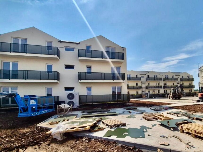 Construction site with beige apartment buildings and multiple balconies, debris and pallets on the ground in the foreground.
