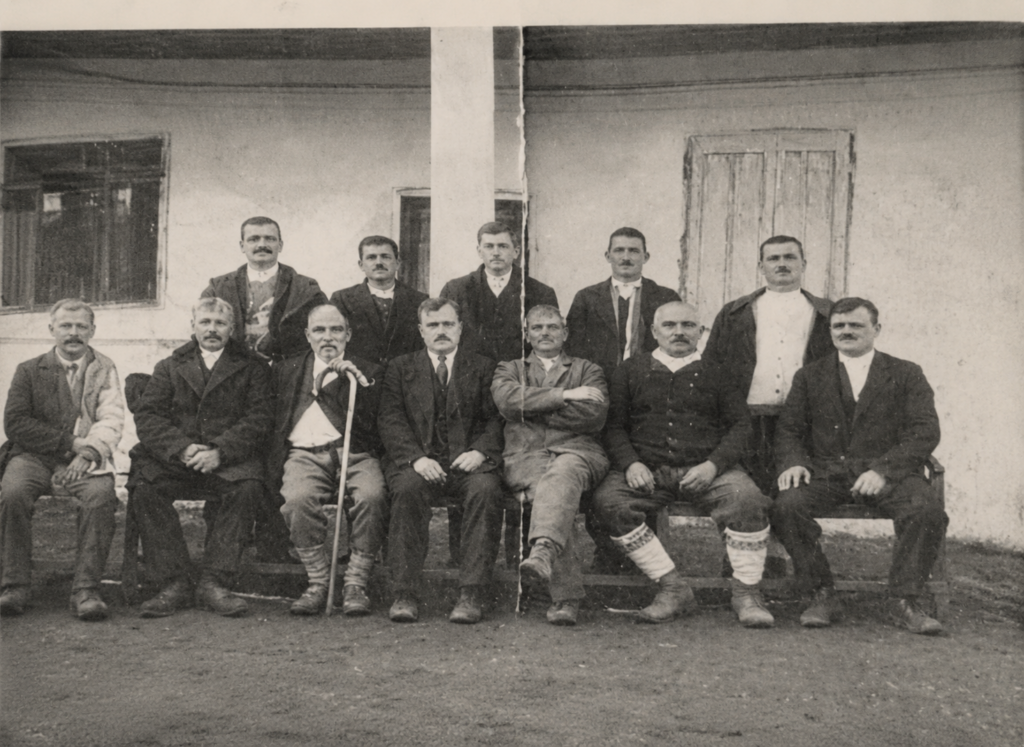 Group of twelve men in early 20th‑century clothing posing for a formal photograph outside a building, some seated on benches and others standing behind them.