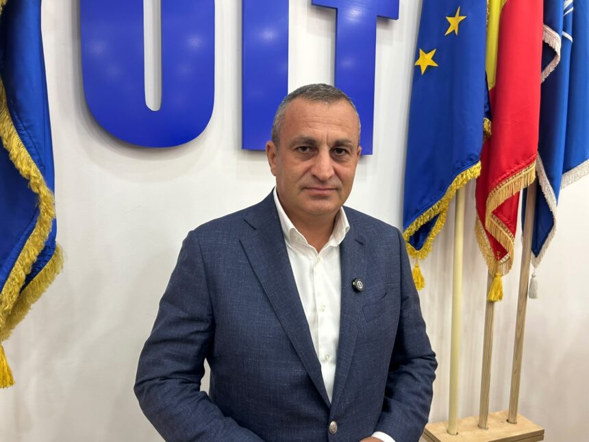 Man in a navy suit and white shirt sits in front of blue flags and gold-fringed banners in a formal setting.