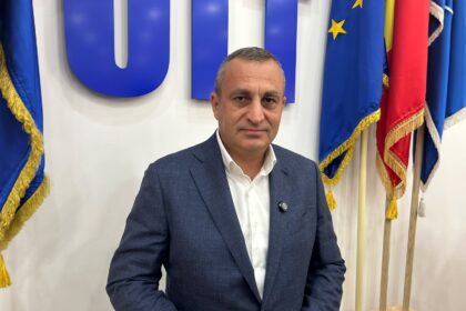 Professional portrait of a man in a dark blue suit and white shirt, standing in front of blue and European flags behind him.