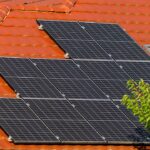 Solar panels mounted on a red tiled roof beside a leafy tree nearby.