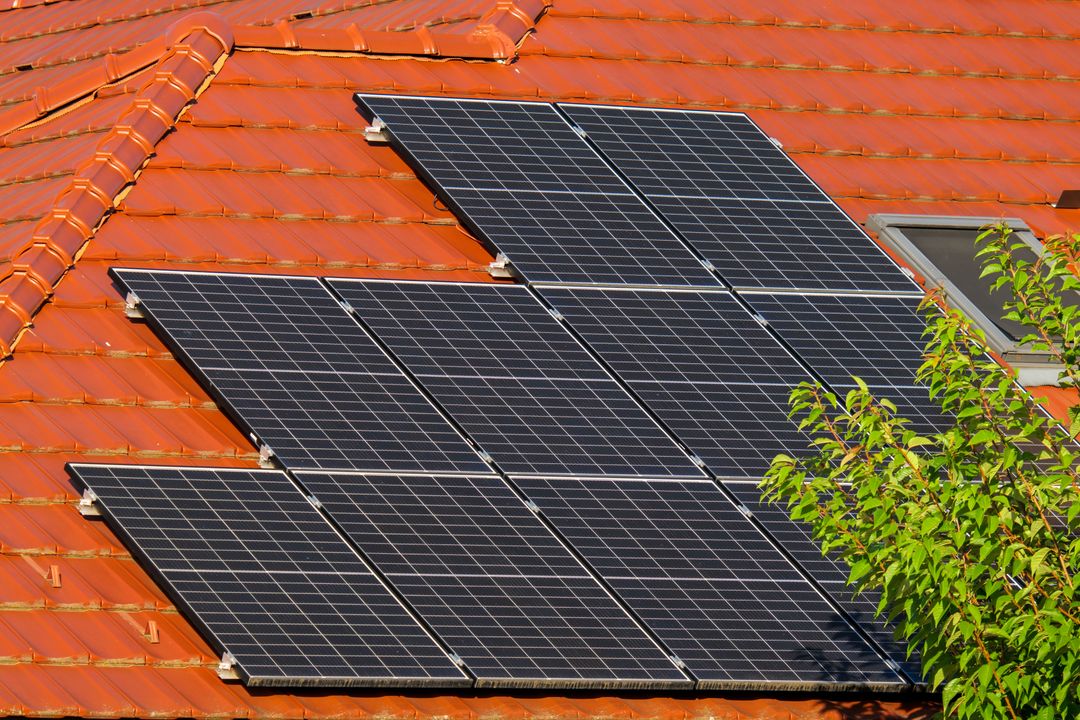 Solar panels mounted on a red tiled roof beside a leafy tree nearby.