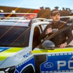 Policeman in tactical vest stands beside an open patrol car door, holding a radio, with blue and red police lights on the roof behind him, on a sunny day.