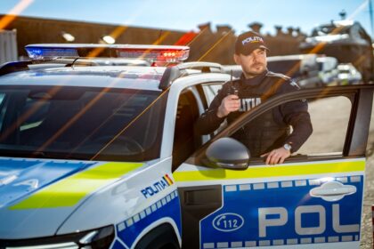 Policeman in tactical vest stands beside an open patrol car door, holding a radio, with blue and red police lights on the roof behind him, on a sunny day.