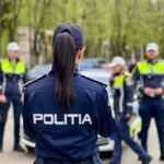 Back view of a police officer in a dark uniform with 'POLITIA' on the back, standing in a street with other officers and a police car in the background.