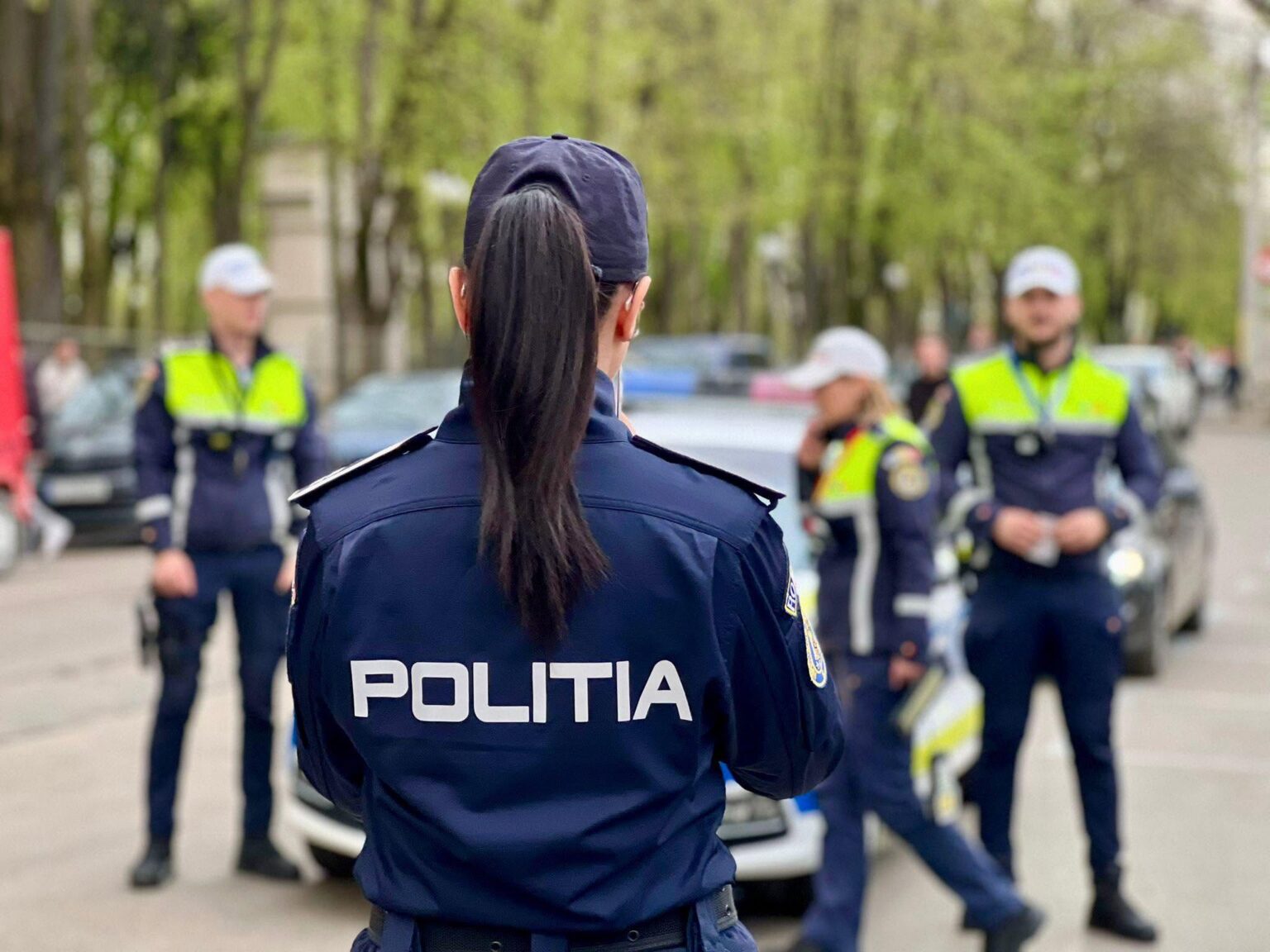 Back view of a police officer in a dark uniform with 'POLITIA' on the back, standing in a street with other officers and a police car in the background.