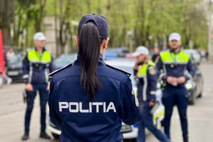 Back view of a police officer in a dark uniform with 'POLITIA' on the back, standing in a street with other officers and a police car in the background.