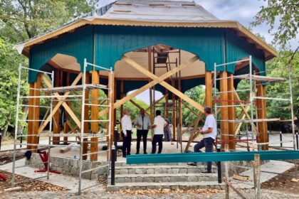 Under-construction wooden pavilion with teal siding in a park; scaffolding surrounds the structure and several workers stand nearby.